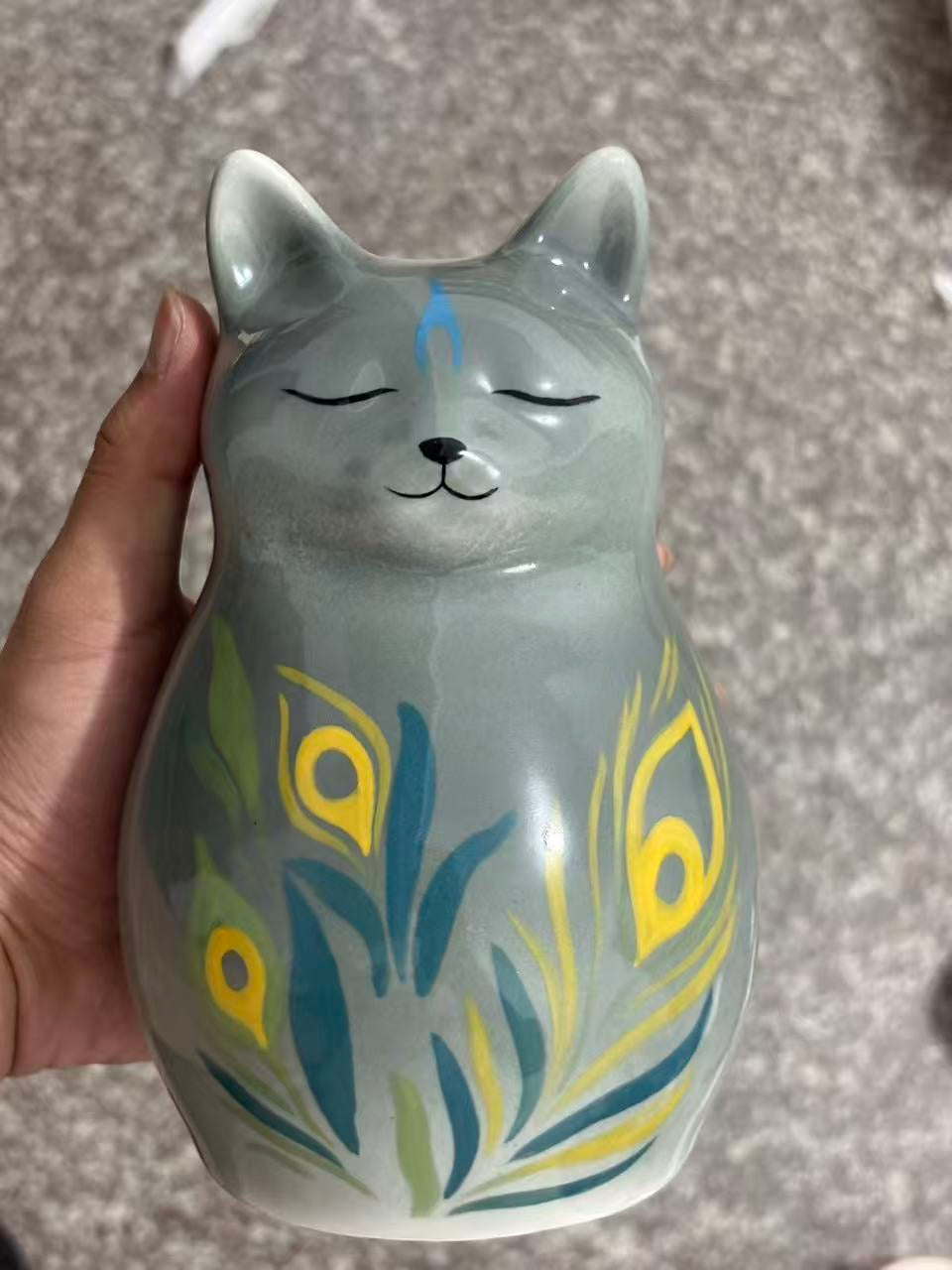 Grey cat urn with closed eyes and long blue, green and yellow decorative leaves across its chest, held in a hand.
