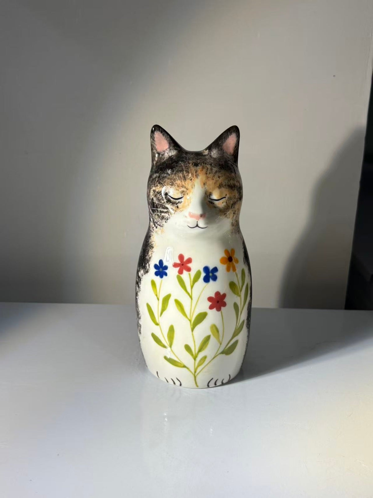 Tabby cat urn with closed eyes, small blue orange red and yellow flowers, green leaves and a painted name.