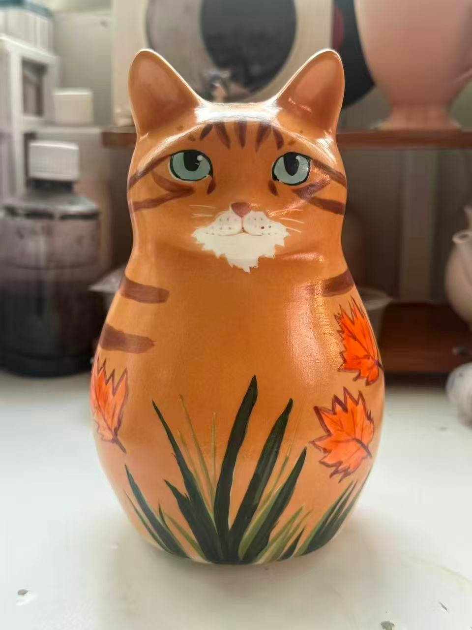 Ginger tabby cat shaped ceramic with orange flowers and long green leaves.